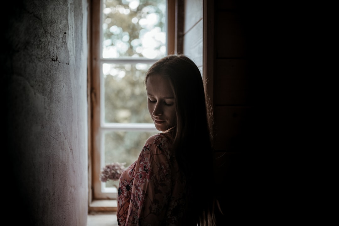 Person stands inside their home, looking out the window with a contemplative or anxious expression, symbolizing the feelin...